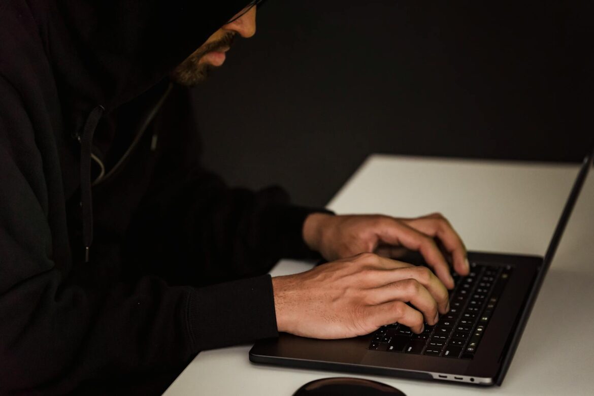 A person in a hoodie working on a laptop in a dimly lit room, representing cybersecurity themes.