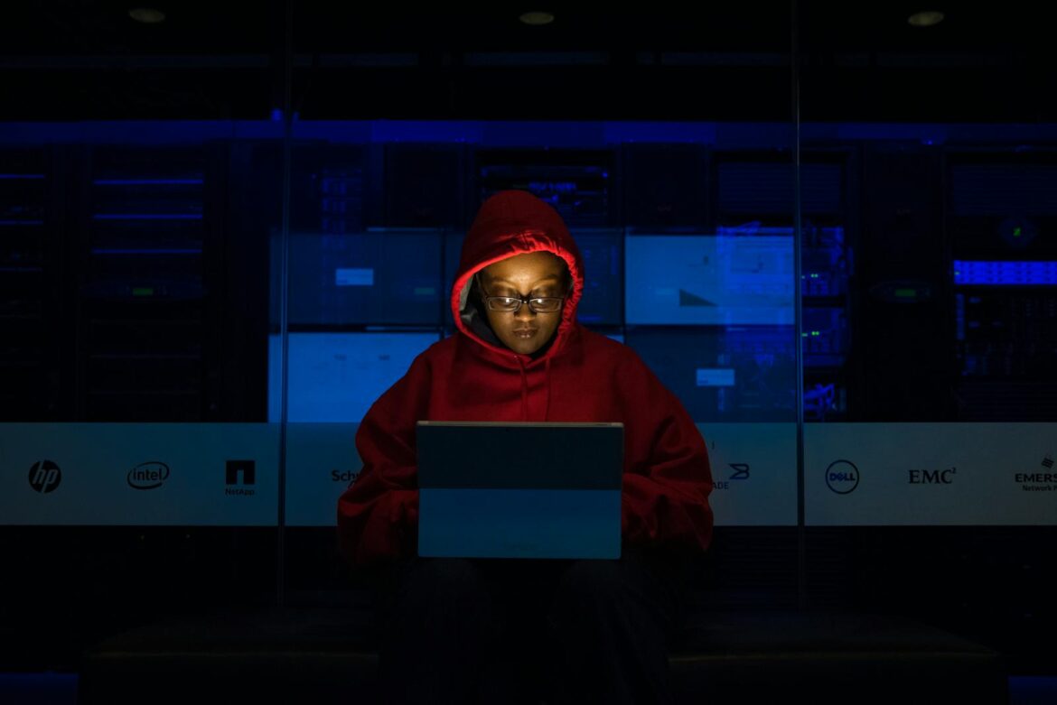 Focused woman in a red hoodie using a laptop in a dimly lit room with technology brands displayed.